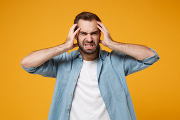 Displeased exhausted young bearded man in casual blue shirt posing isolated on yellow orange background, studio portrait. People emotions lifestyle concept. Mock up copy space. Putting hands on head.