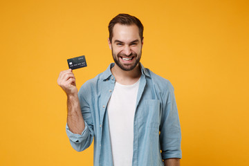 Smiling young man in casual blue shirt posing isolated on yellow orange wall background, studio portrait. People sincere emotions lifestyle concept. Mock up copy space. Hold in hand credit bank card. - obrazy, fototapety, plakaty