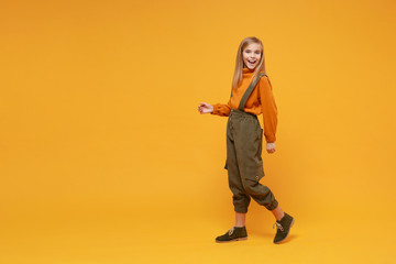 Fototapeta na wymiar Laughing little blonde kid girl 12-13 years old in turtleneck jumpsuit isolated on orange yellow background children studio portrait. Childhood lifestyle concept. Mock up copy space. Looking camera.