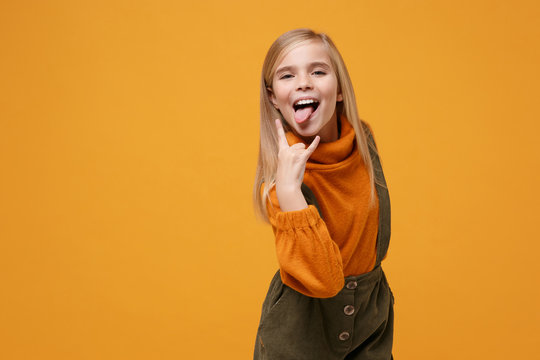 Funny little kid girl 12-13 years old in turtleneck jumpsuit isolated on orange yellow wall background. Childhood lifestyle concept. Mock up copy space. Showing tongue depicting heavy metal rock sign.