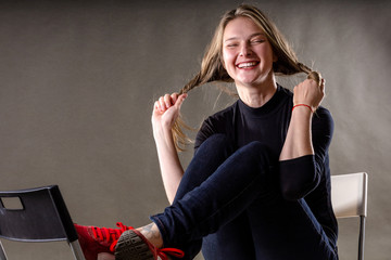 smiling young blonde girl sitting on a chair and playing with hair on a gray background