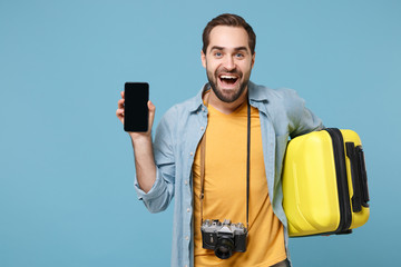 Excited traveler tourist man in casual clothes with photo camera isolated on blue background. Passenger traveling abroad on weekends. Air flight journey. Hold suitcase, mobile phone with empty screen.