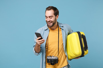 Cheerful traveler tourist man in casual clothes with photo camera isolated on blue background. Male passenger traveling abroad on weekends. Air flight journey concept Hold suitcase using mobile phone. - obrazy, fototapety, plakaty