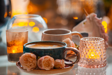 A cup of coffee on a saucer is decorated with roses next to candles in beautiful candlesticks on a glass table