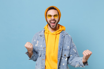 Happy young hipster guy in fashion jeans denim clothes posing isolated on pastel blue background studio portrait. People sincere emotions lifestyle concept. Mock up copy space. Doing winner gesture.