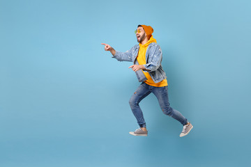 Side view of excited young hipster guy in fashion jeans denim clothes posing isolated on pastel blue background. People lifestyle concept. Mock up copy space. Jumping pointing index fingers aside.