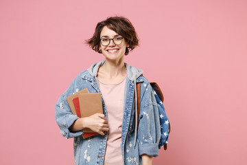 Smiling young woman student in denim clothes, eyeglasses, backpack posing isolated on pastel pink background. Education in high school university college concept. Mock up copy space. Holding books.
