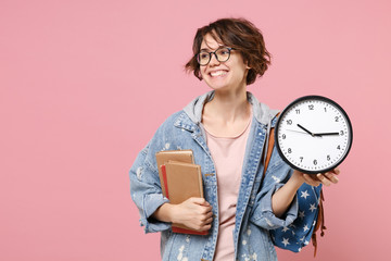Pretty young woman student in denim clothes eyeglasses, backpack posing isolated on pastel pink background. Education in high school university college concept. Mock up copy space. Hold books clock.