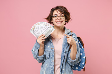 Smiling girl student in denim clothes glasses backpack isolated on pastel pink background. Education in school university college concept. Point index finger on fan of cash money in dollar banknotes. - obrazy, fototapety, plakaty