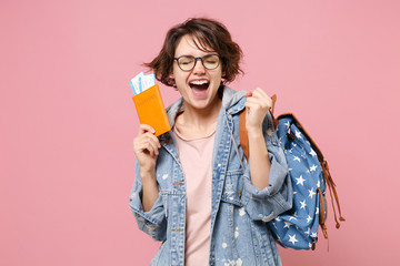 Joyful young woman student in denim clothes glasses backpack isolated on pink background. Education in high school university college concept. Hold passport boarding pass tickets doing winner gesture.
