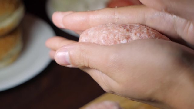 Slow motion view of hands shaping minced beef into a cutlet for a burger with a bun and tomatoes with lettuce in the background