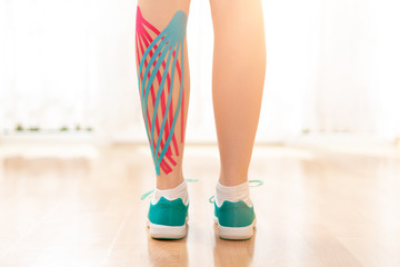 Female patient with kinesio tape on her calf standing in front of a window, rear view leg close up....