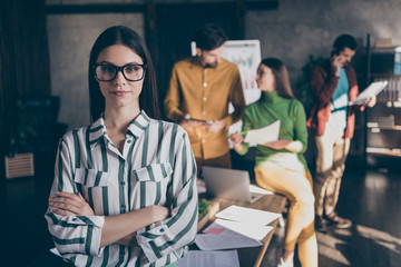 Photo of serious confident woman standing with arms crossed wearing spectacles in striped shirt formalwear on background of her coworkers discussing startup strategy