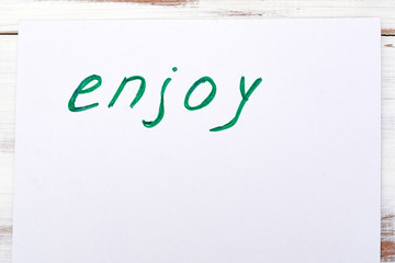 Word enjoy written on a white piece of paper on a light background.