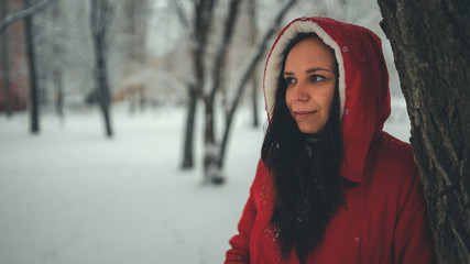 Portrait of young woman in red jacket and hood in winter season. Beautiful lady stands near tree and looks away. Fluffy snow envelops everything around.