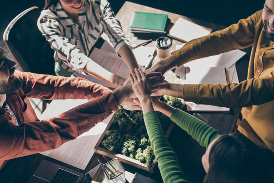 Cropped top above high angle view of four nice cheerful professional businesspeople wearing casual formalwear putting hands together over desk at modern interior workplace station