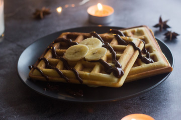 Viennese waffles on a plate with banana and chocolate. In the background are lights and candles. Romantic picture