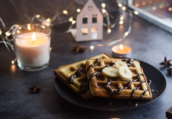 Viennese waffles on a plate with banana and chocolate. In the background are lights and candles. Romantic picture