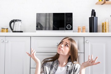 Fototapeta na wymiar confused and attractive woman looking at microwave and doing shrug gesture in kitchen