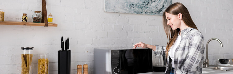 panoramic shot of smiling woman looking at microwave in kitchen