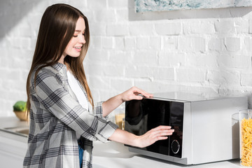 Fototapeta na wymiar smiling and attractive woman in shirt using microwave in kitchen