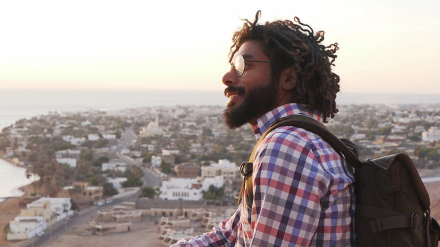 smiling adult traveler with dreadlocks sitting on a cliff at sunrise