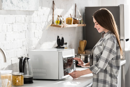 side view of smiling woman in shirt using microwave in kitchen