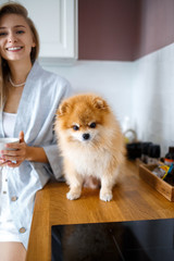 Happy blonde woman is drinking tea in the kitchen, next to her is her Pomeranian breed dog. The friendship of people and animals.