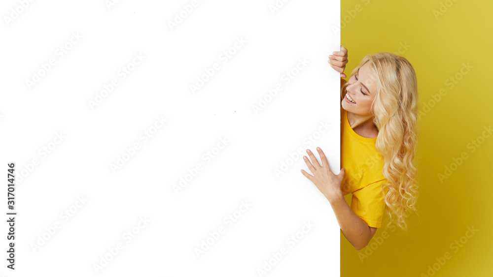 Wall mural young happy woman peeks out from behind a white banner on a yellow background. point to an empty bla