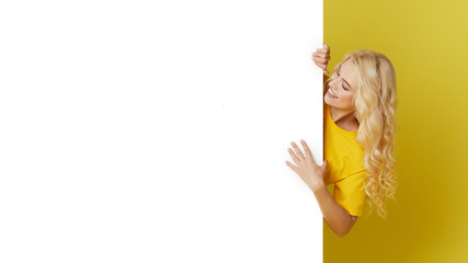 Young happy woman peeks out from behind a white banner on a yellow background. Point to an empty blank on a form, a copy space for text. Horizontal shot - obrazy, fototapety, plakaty