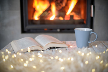 Open book and cup of tea near burning fireplace. Hygge concept
