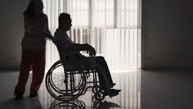 Silhouette of old woman pushing her husband on wheelchair after recovering from stroke disease at home. Shot in 4k resolution