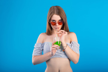 Young woman with green drink in drinking glass and glasses in white striped swimsuit lingerie isolated on blue background