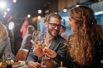 Cute caucasian couple sitting in restaurant and cheering with wine. In background are their friends. - obrazy, fototapety, plakaty