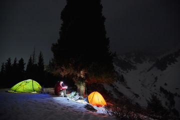 Man sits near a tents at night in the winter in the mountains