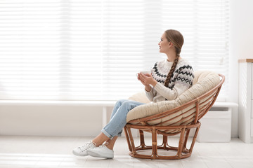 Young woman with hot drink in papasan chair near window at home