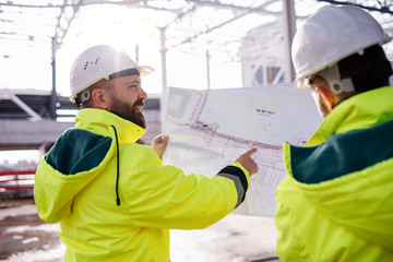 Men engineers standing outdoors on construction site, holding blueprints.