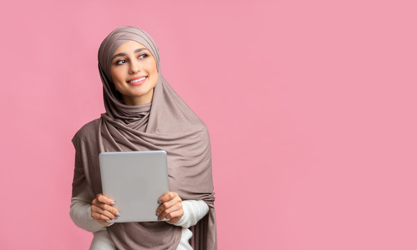 Smiling islamic woman in headscarf holding digital tablet and looking aside