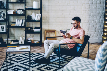 Caucasian hipster guy resting in comfortable flat with loft interior and using home free internet for installing application for communicate with friends in social network via digital tablet