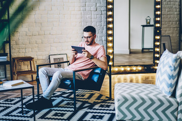 Fototapeta premium Young male blogger resting at own apartment with stylish comfortable interior and using modern touch pad for watching video online on web page, hipster guy relaxing at home with new digital tablet