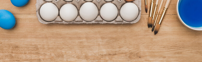 top view of blue watercolor paint in bowl near chicken eggs and paintbrushes on wooden table, panoramic shot