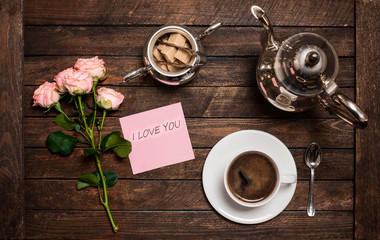 Roses and coffee. Cup of coffee, vintage silver coffeepot,  sugar bowl and a bouquet of roses on a wooden table. Morning coffee and note I love you.