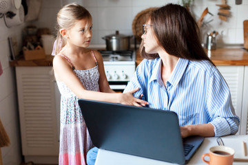 Working mother in home office. Woman and child girl using laptop. Daughter needs attention from irritated, exhausted, stressed mom. Freelancer workplace in kitchen. Female business. Lifestyle moment.