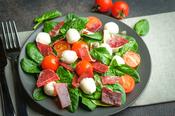 Salad with prosciutto, mozzarella, fresh vegetables on a dark plate on a black background.