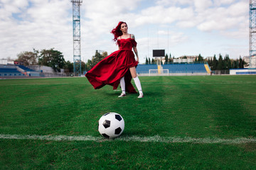 Luxurious woman with red hair and in a red dress plays on the football field.