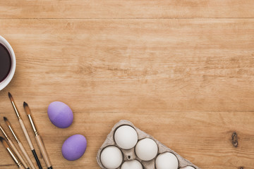 top view of watercolor purple paint in bowl near chicken eggs and paintbrushes on wooden table