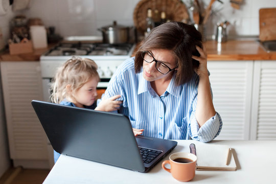 Working mother in home office. Unhappy woman and child using laptop. Sad and angry daughter needs attention from busy exhausted mom. Freelancer workplace in kitchen. Female business. Lifestyle moment.