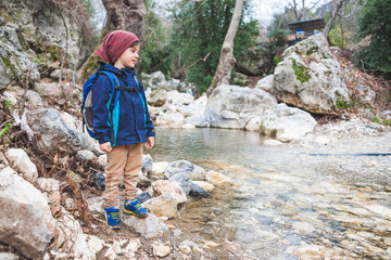 A child with a backpack stands near a mountain river.