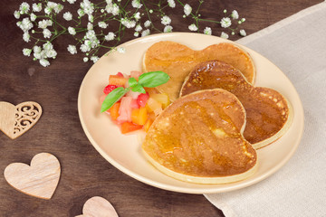 Homemade heart shaped pancakes with fruit chutney salad and honey on a beige plate on a wooden table. Rustic style breakfast or brunch for Valentine's Day. Top view, copyspace, closeup