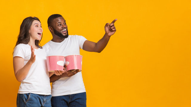 Excited interracial couple holding popcorn backets and pointing at copy space